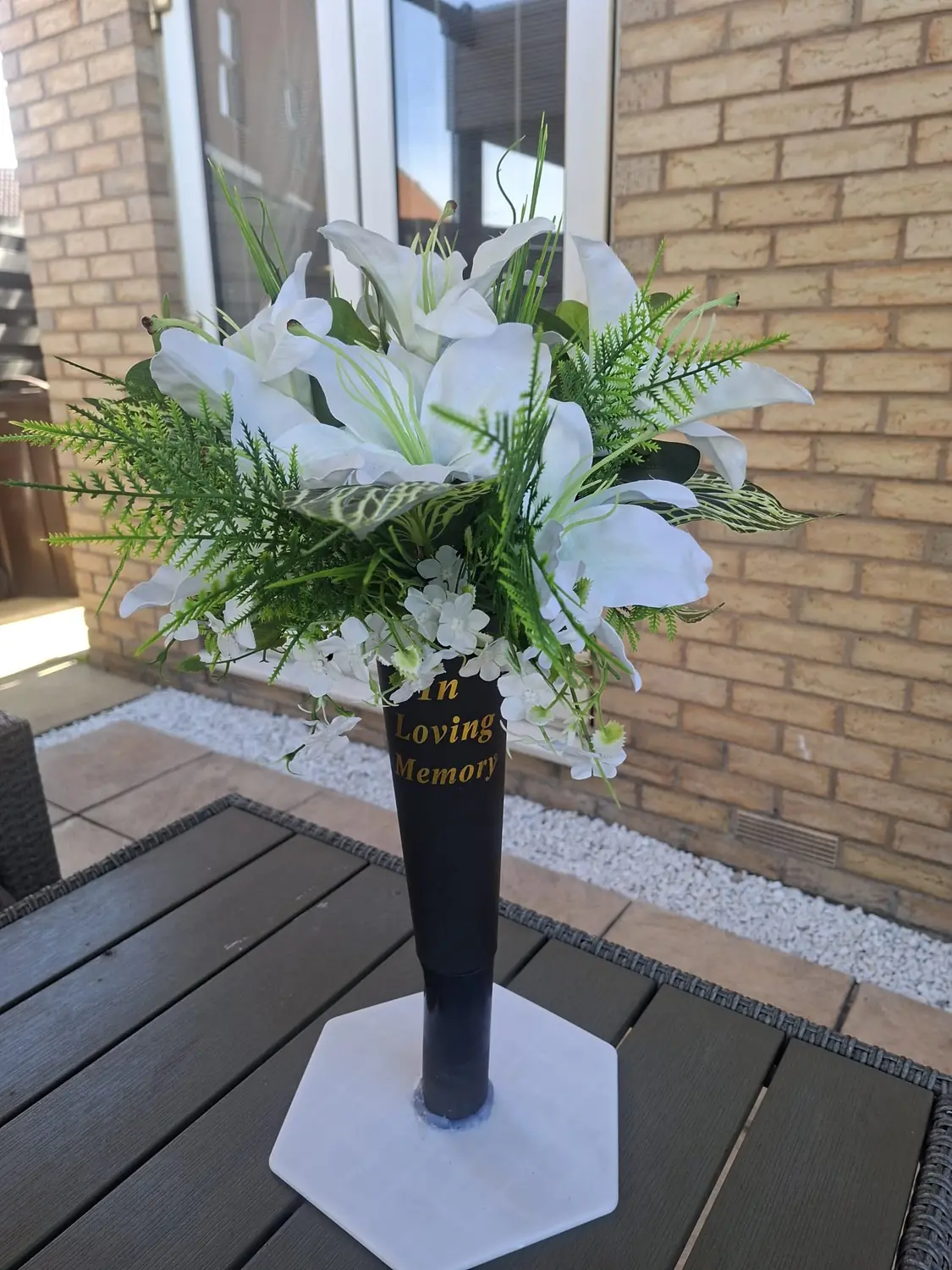 A graveside vase of white lilies, white blossom and green ferns in a black In Loving Memory holder.